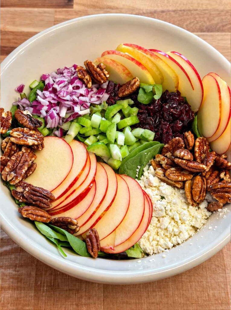 A bowl of salad with apples, feta cheese, maple pecans, red onions, celery and greens