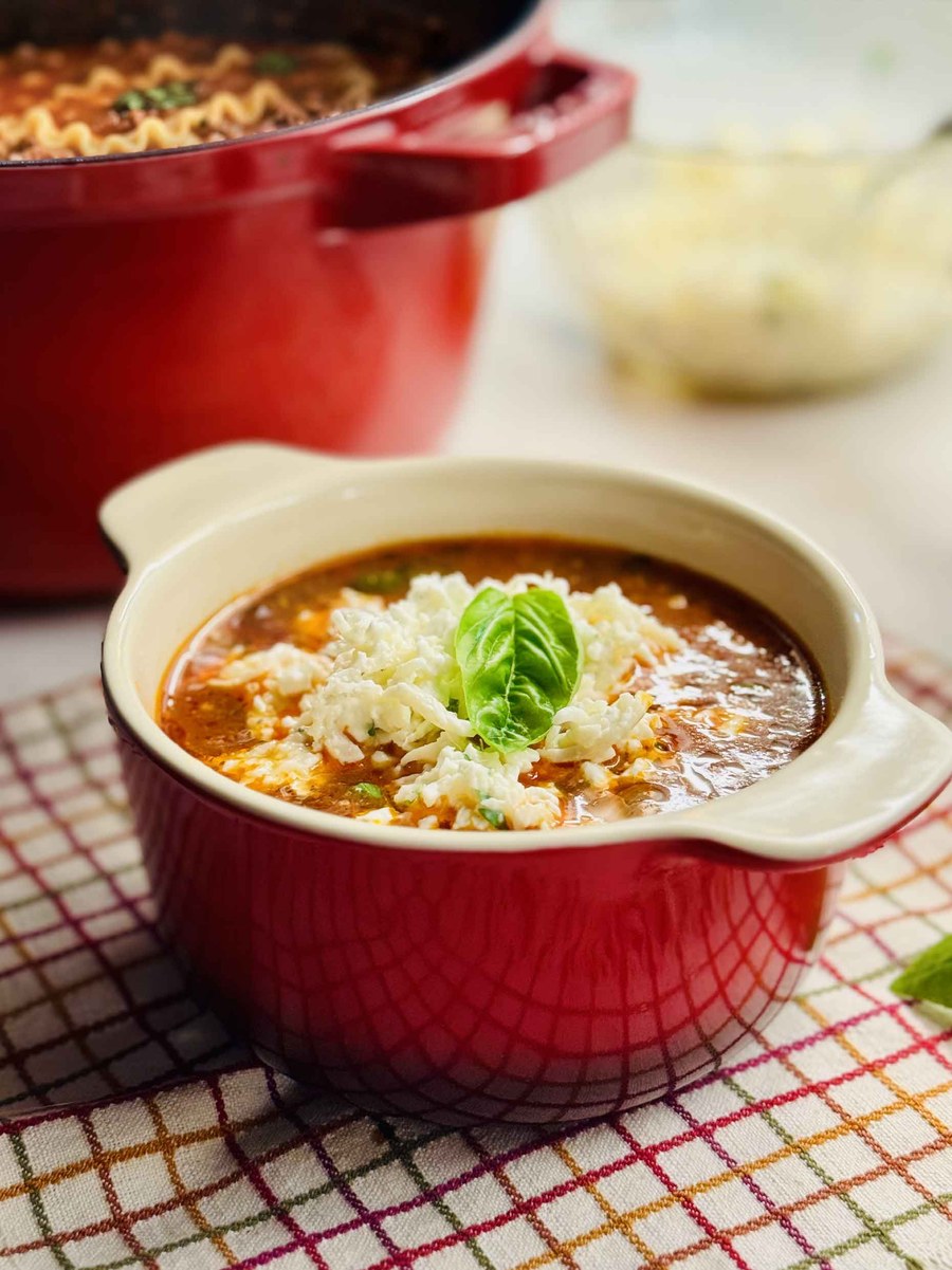 A red bowl of lasagna soup topped with a mound of three-cheese topping and a fresh basil leaf, with a red Dutch oven behind it