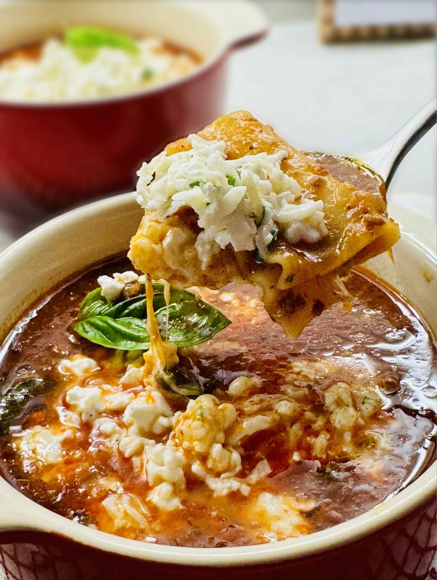 A spoon lifting a lasagna noodle with melted mozzarella stretching down into a bowl of lasagna soup with cheese and fresh basil