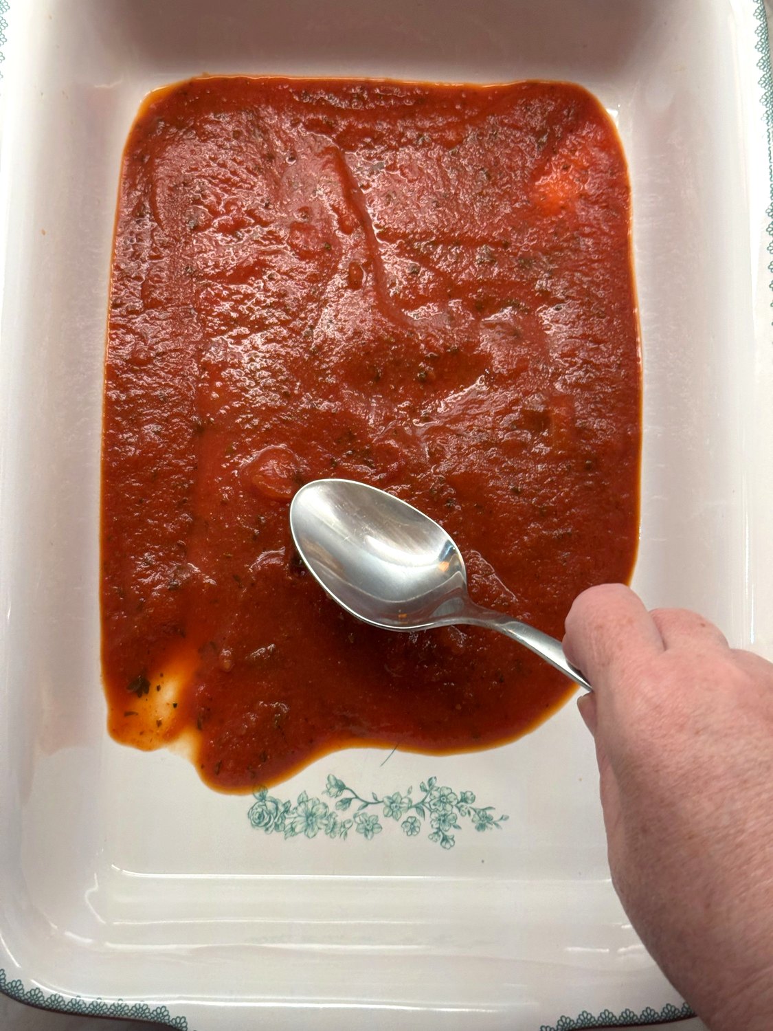 Marinara sauce being spread over the bottom of a white baking dish with a spoon