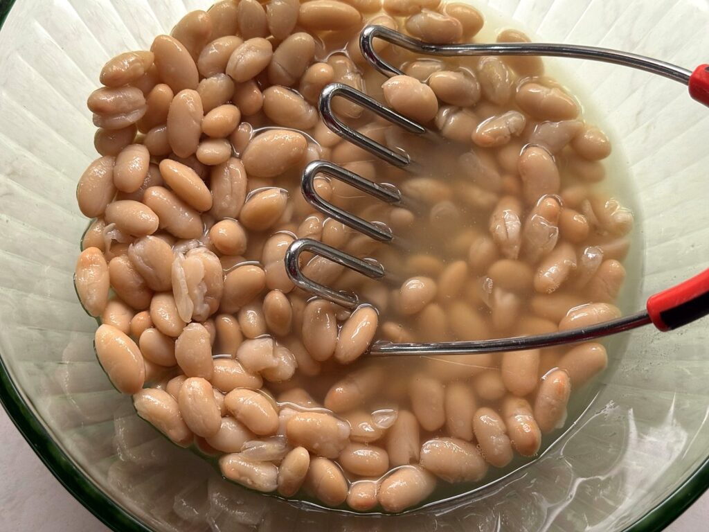 A potato masher in a glass bowl of great northern beans and broth
