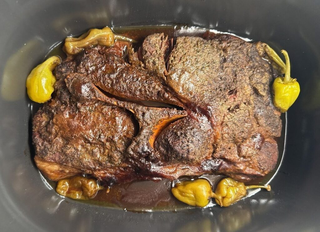 A cooked Mississippi pot roast with a dark seasoned crust and pepperoncini, sitting in its juices in the slow cooker