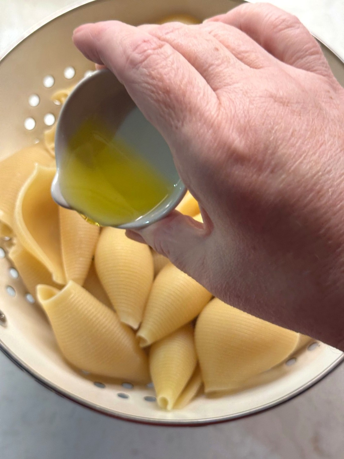 Olive oil being drizzled over drained jumbo shells in a colander