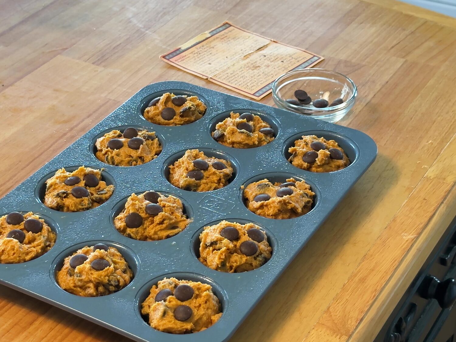 Twelve unbaked pumpkin chocolate chip muffins in a muffin pan, topped with chocolate, ready for the oven