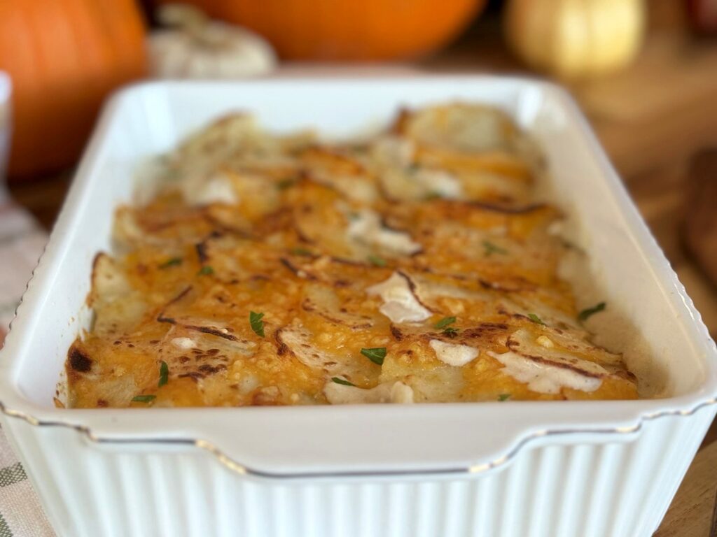 Cheesy scalloped potatoes with golden, shingled potato slices and parsley in a white baking dish, with pumpkins in the background