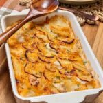 A side dish of scalloped potatoes with shingled potatoes in a white baking dish and wooden spoon