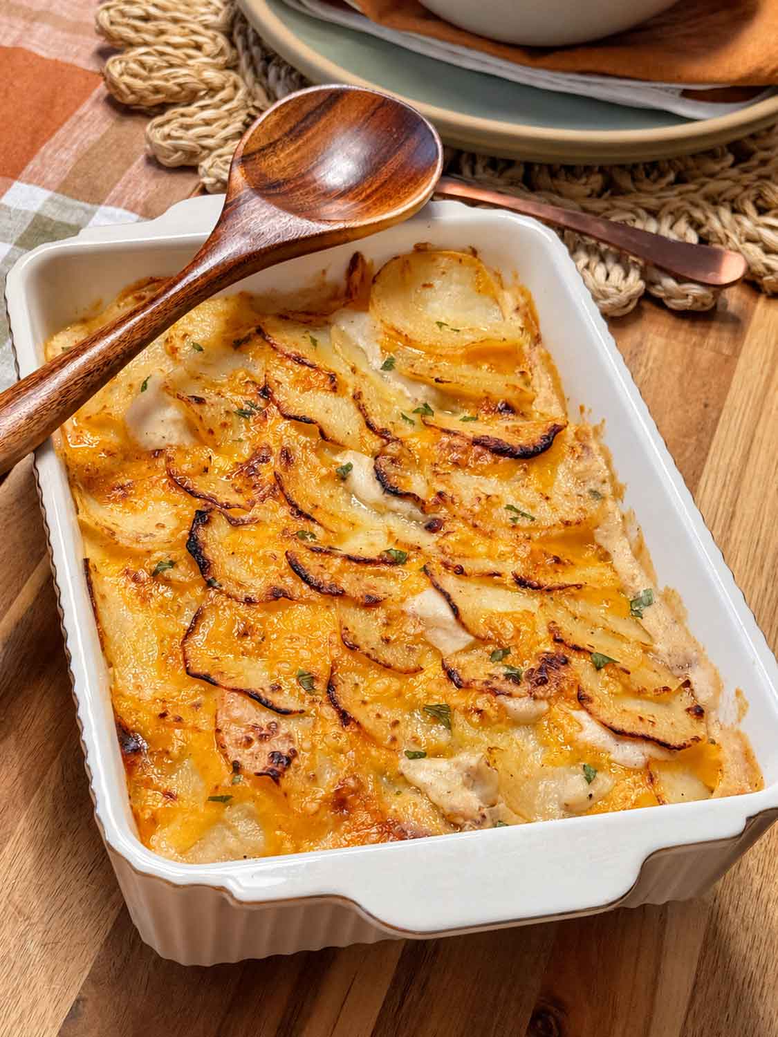 A side dish of scalloped potatoes with shingled potatoes in a white baking dish and wooden spoon