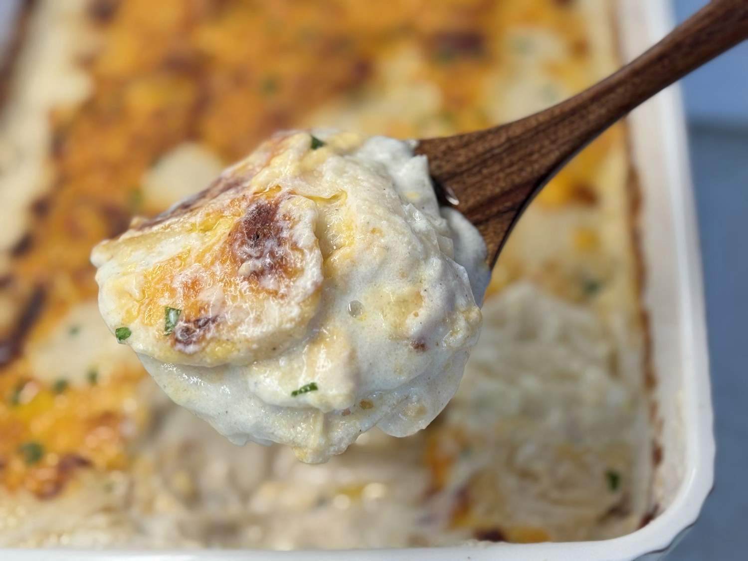 Wooden spoon lifting a creamy scoop of saucy scalloped potatoes out of the baking dish