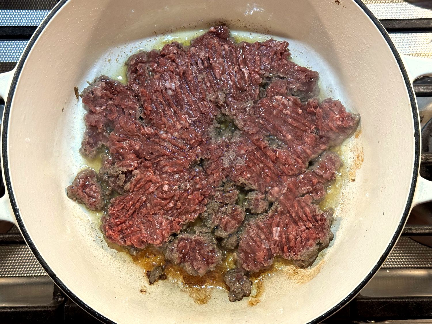 Raw ground venison pressed into an even layer in olive oil in a white enameled Dutch oven