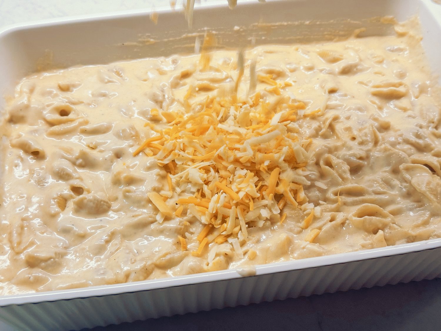 Saucy pasta shells in cheese sauce in a white baking dish, with a pile of shredded cheddar and Colby Jack on top