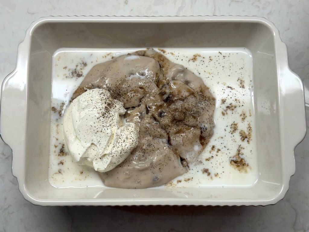 Cream of mushroom soup, sour cream, milk, and black pepper in a white baking dish before stirring