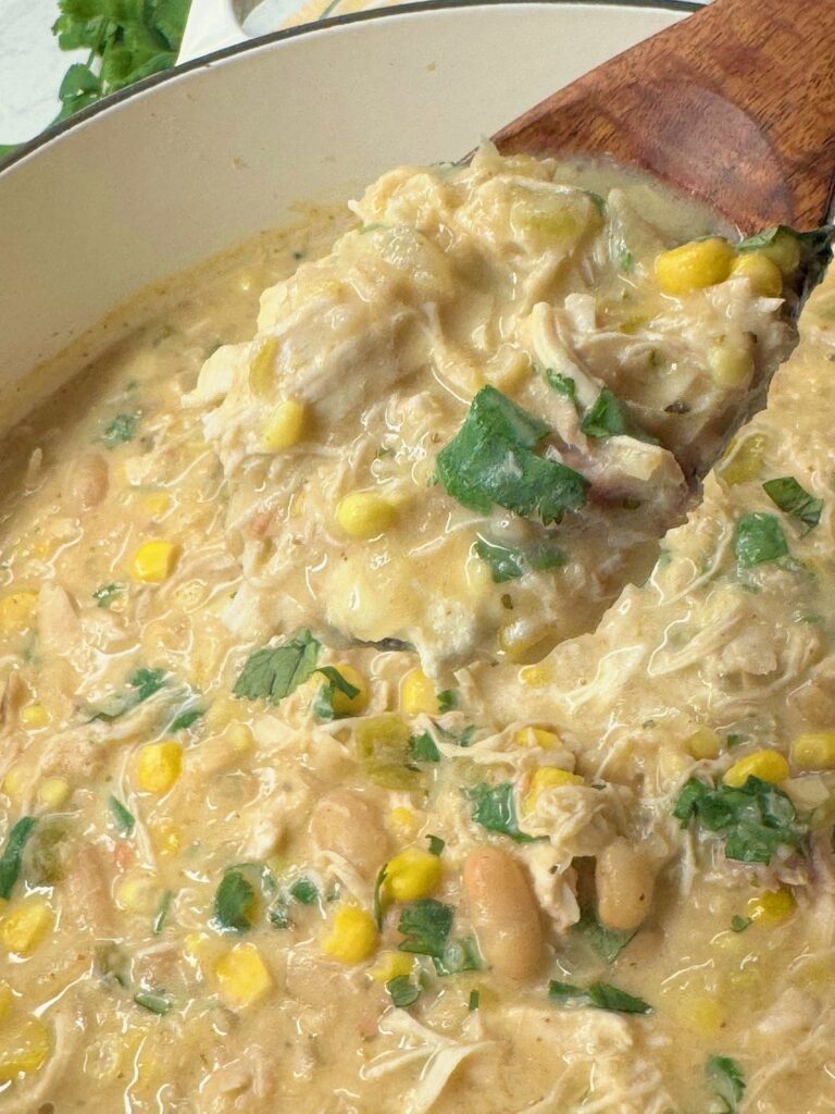 A wooden spoon lifting thick white chicken chili with chicken, beans, and corn from the pot