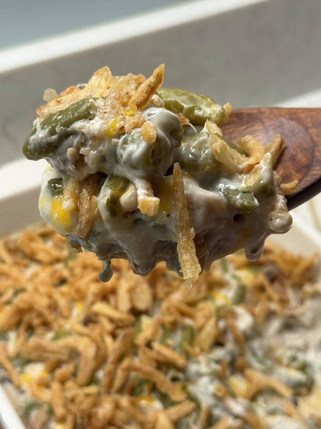 A wooden spoon lifting a creamy spoonful of green bean casserole with crispy onions and melted cheddar