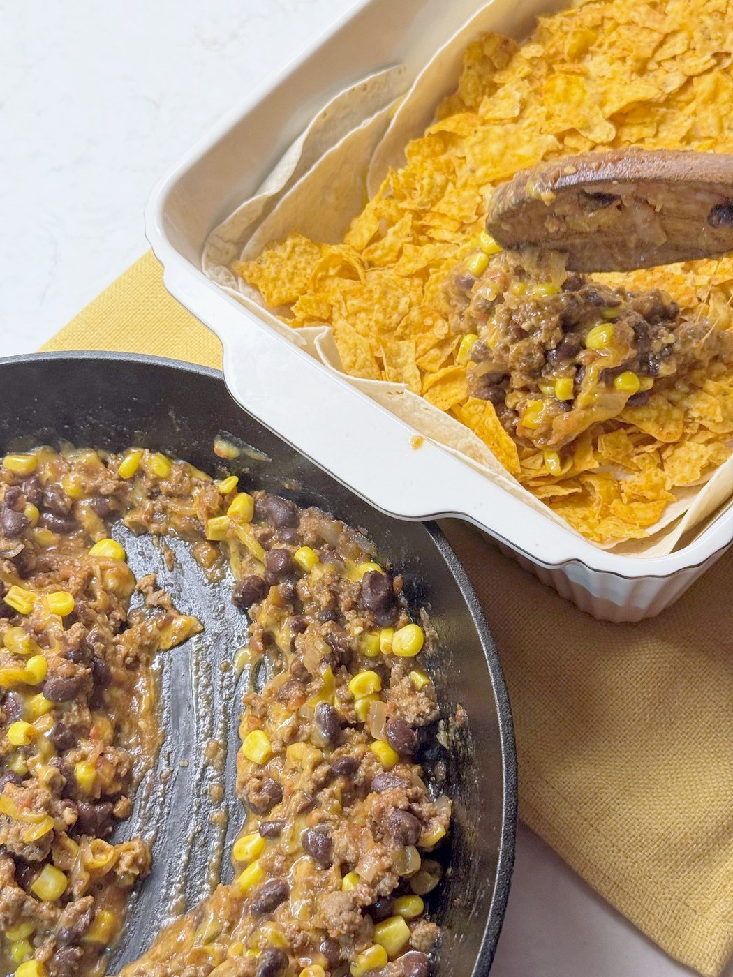 A wooden spoon spooning taco beef filling from a cast iron skillet over the Doritos layer