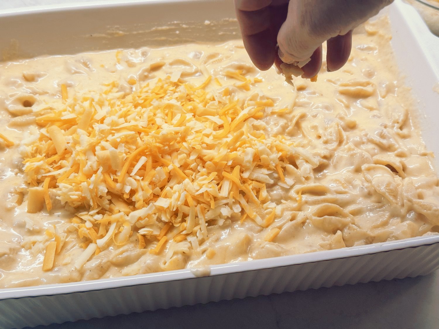 A hand sprinkling shredded cheese over saucy mac and cheese in a white baking dish