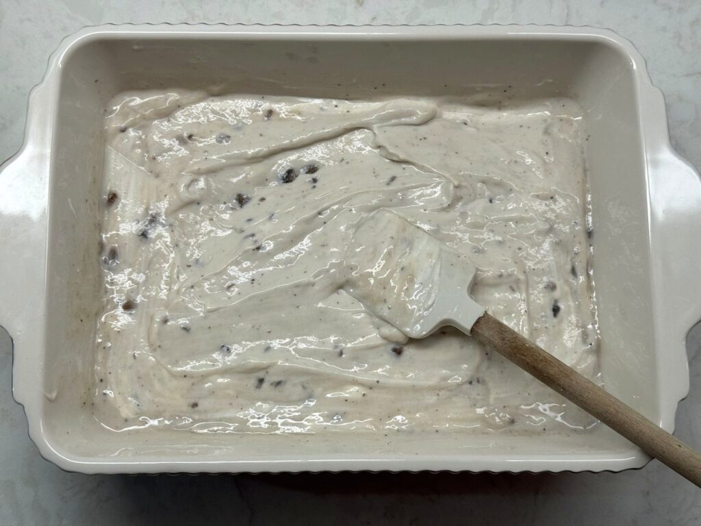 A spatula stirring a smooth, creamy sauce in a white baking dish