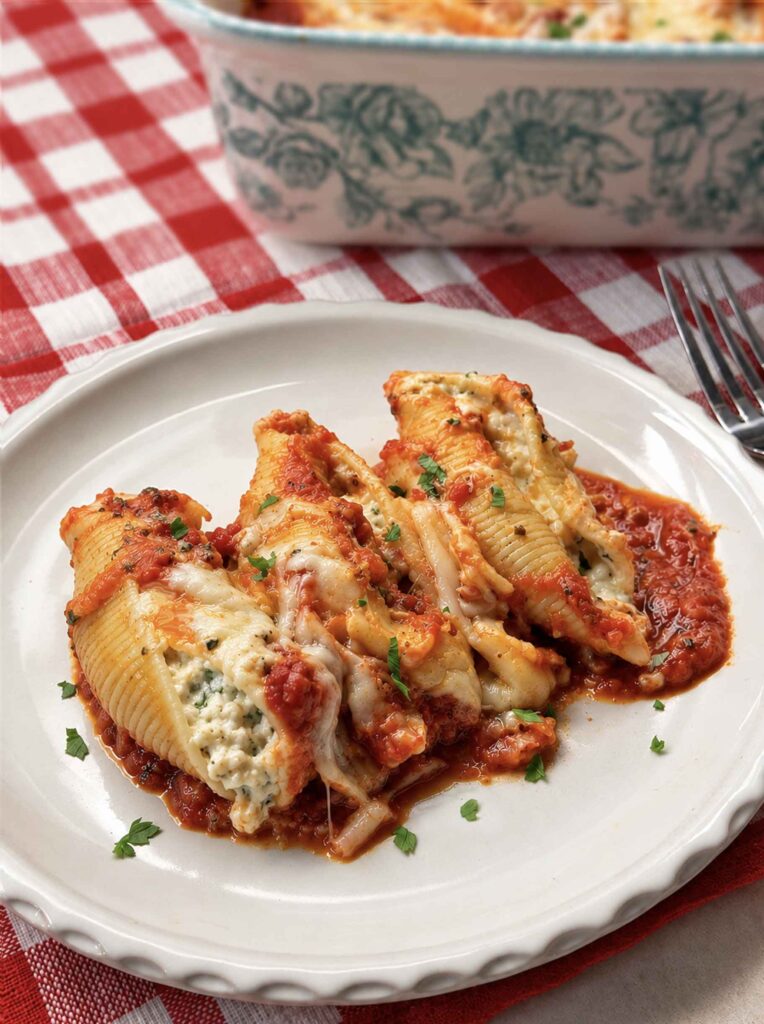 Three stuffed shells on a white plate, with the baking dish filled with shells behind it. 