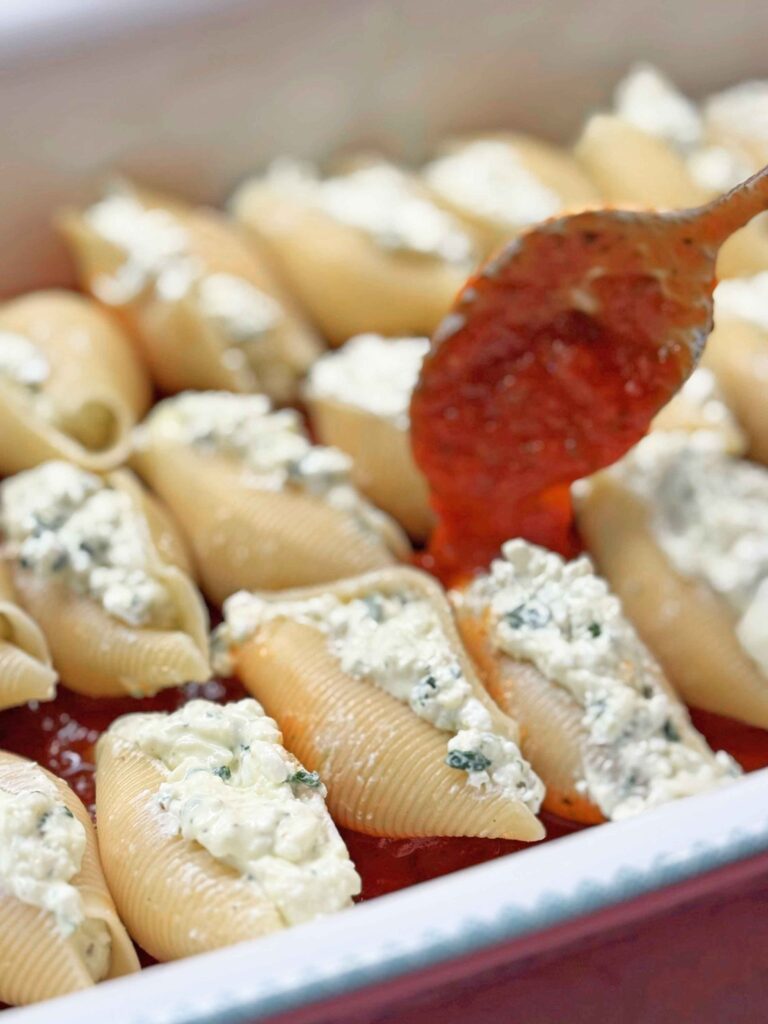 Marinara sauce being spooned over filled jumbo shells in a baking dish