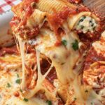 A baked stuffed shell lifted from the pan with a wooden spatula, trailing strings of melted mozzarella
