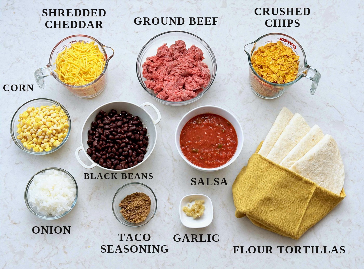 Labeled ingredients for taco casserole: shredded cheddar, ground beef, crushed Doritos, corn, black beans, salsa, flour tortillas, onion, taco seasoning, and garlic