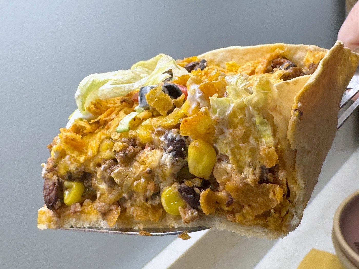 A square of taco casserole on a spatula showing the flour tortilla crust, softened Doritos, beef, beans, corn, and cheese