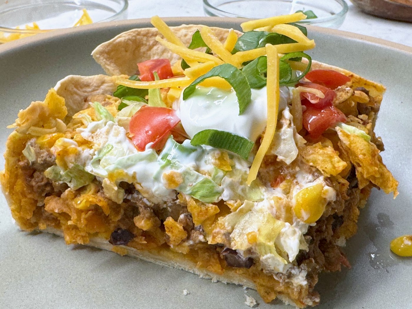 A square of taco casserole on a plate topped with sour cream, tomato, green onion, and shredded cheddar
