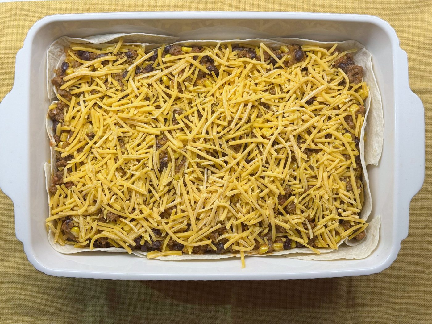 Taco casserole topped with shredded cheddar in a white baking dish, ready to bake