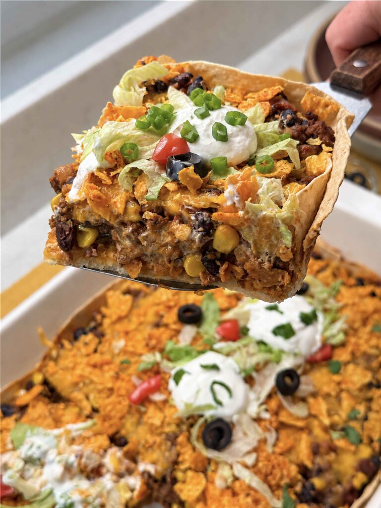 Serving a piece of taco casserole with toppings of sour cream, green onions, tomatoes, and olives on top.