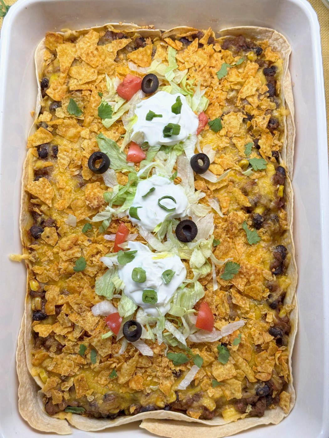 Overhead view of taco casserole in a 9x13 baking dish with a crushed Doritos topping and a stripe of lettuce, tomato, sour cream, green onion, black olives, and cilantro down the middle
