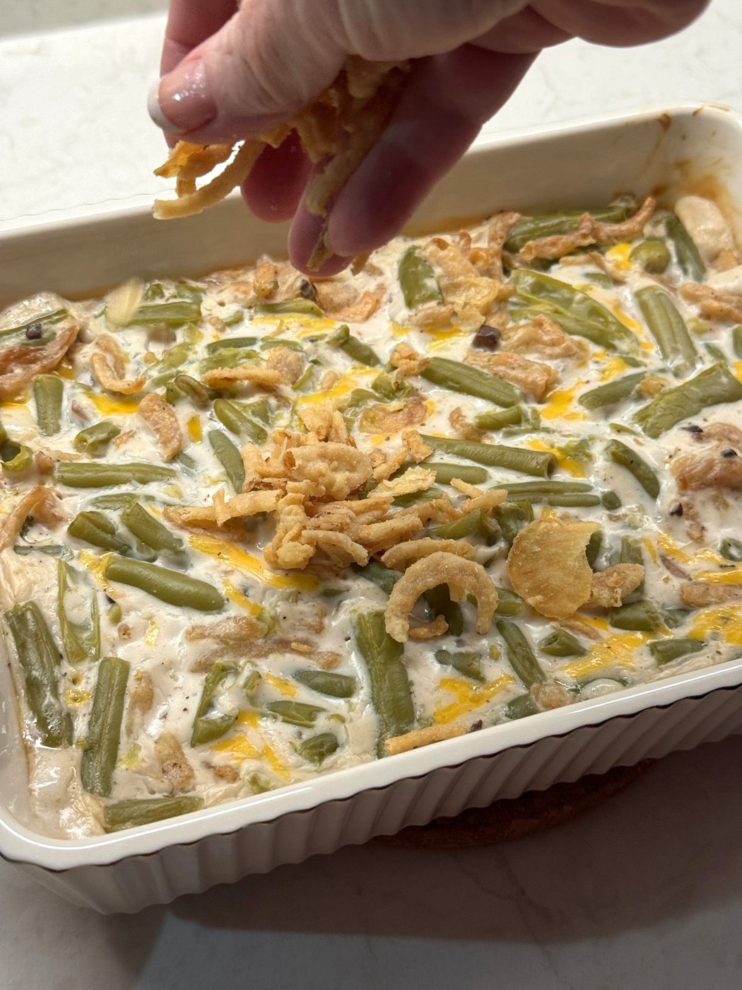 A hand sprinkling fried onions over a hot green bean casserole