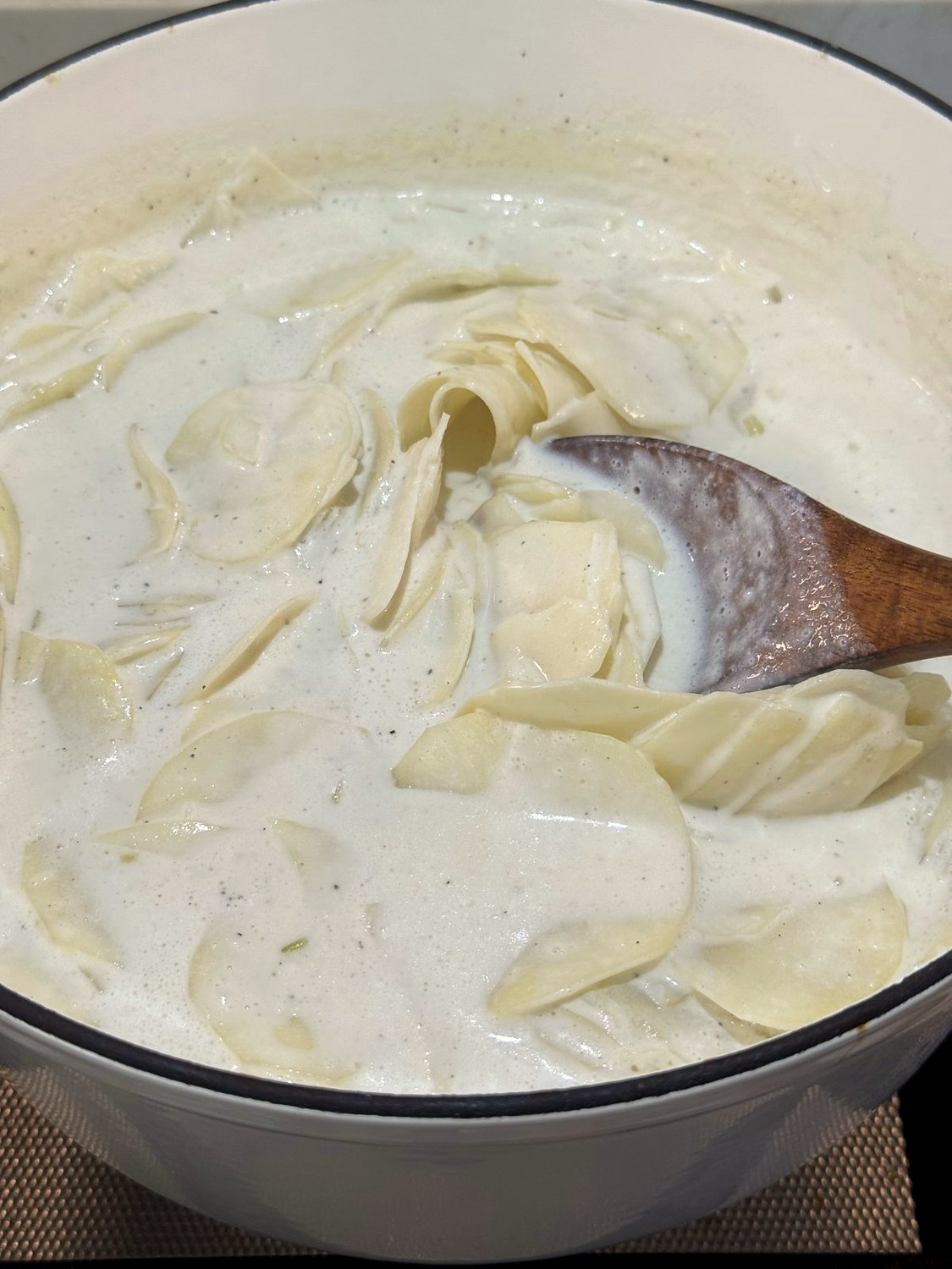Wooden spoon gently turning thin potato slices in thickened cream sauce in a Dutch oven