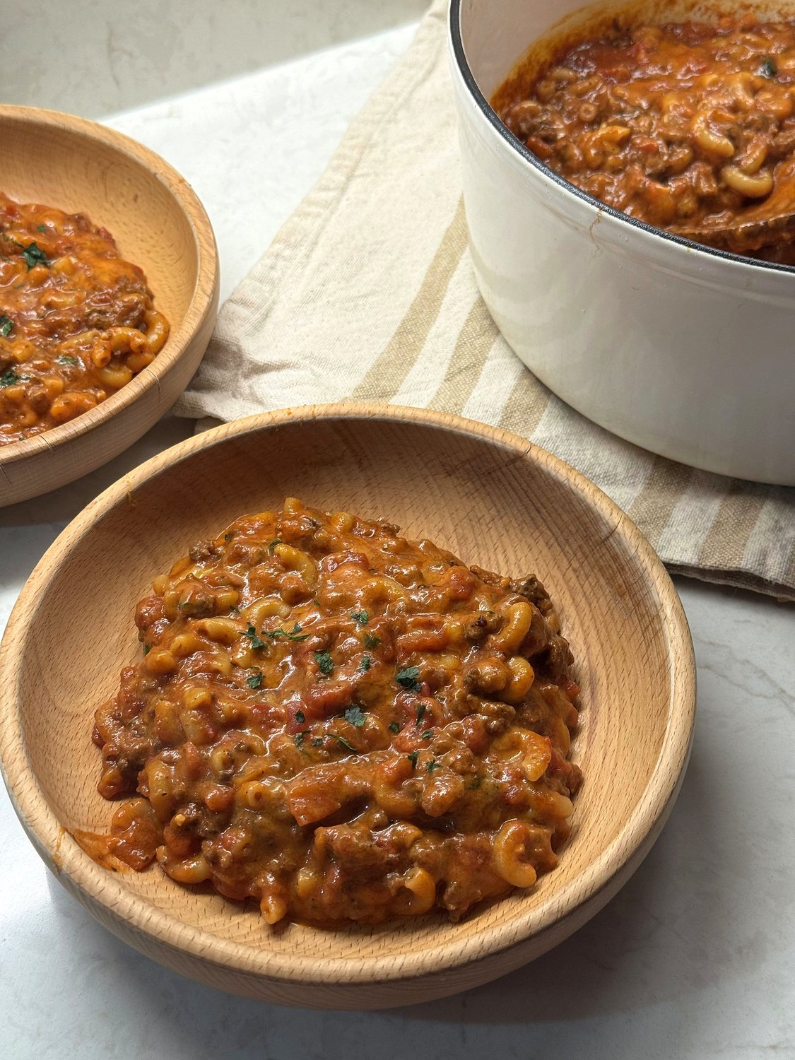 Two wooden bowls of venison goulash beside a white Dutch oven on a striped kitchen towel