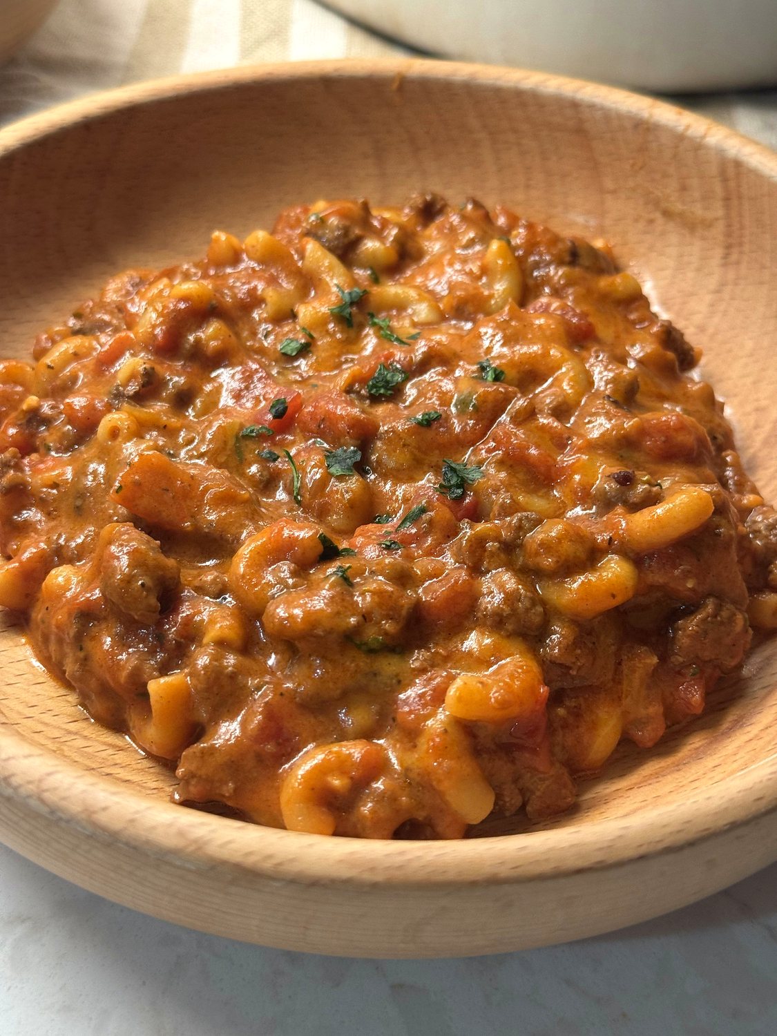 A wooden bowl of cheesy venison goulash with elbow macaroni, ground venison, and tomato sauce, garnished with chopped parsley