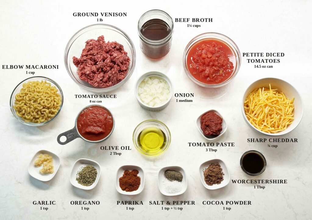Labeled ingredients for venison goulash: ground venison, beef broth, petite diced tomatoes, elbow macaroni, onion, tomato sauce, olive oil, tomato paste, sharp cheddar, garlic, oregano, paprika, salt and pepper, cocoa powder, and Worcestershire