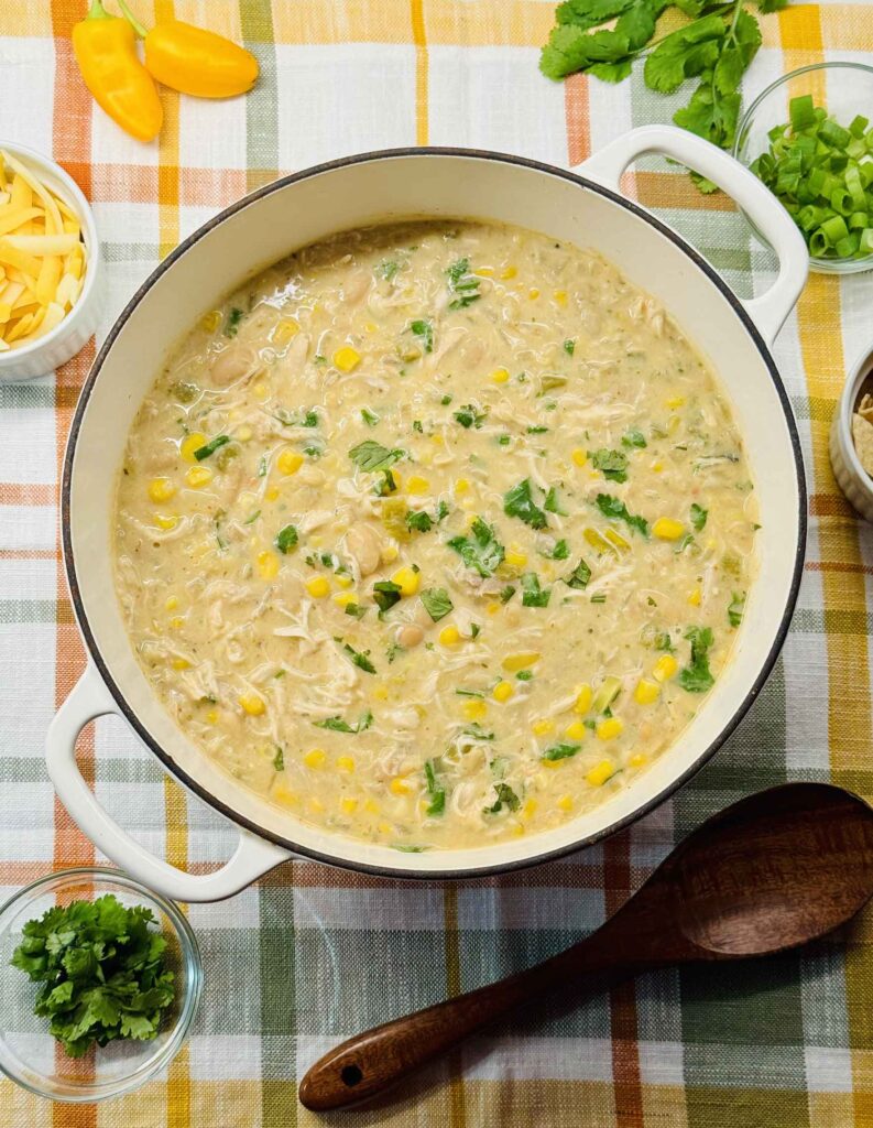 A big pot of white chicken chili with bowls of toppinsg containing with shredded cheddar, sour cream, and cilantro