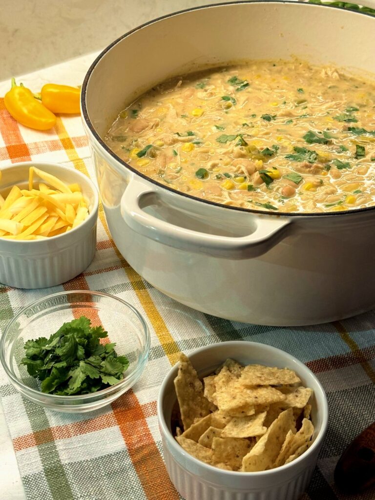 A white Dutch oven of white chicken chili with bowls of shredded cheese, cilantro, and tortilla chips on a plaid towel